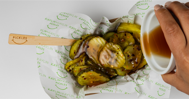 A basket of pickle slices with a wooden spoon sticking out one side that says “pickles.” A ramekin of brown sauce is being poured over the pickles.