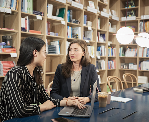 Two professionals having a discussion at a table with laptops and coffee in a modern library or bookstore setting with wooden shelving