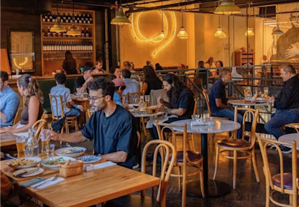 Bustling restaurant interior with warm lighting, neon wall art, and diners seated at wooden tables enjoying meals in a cozy atmosphere